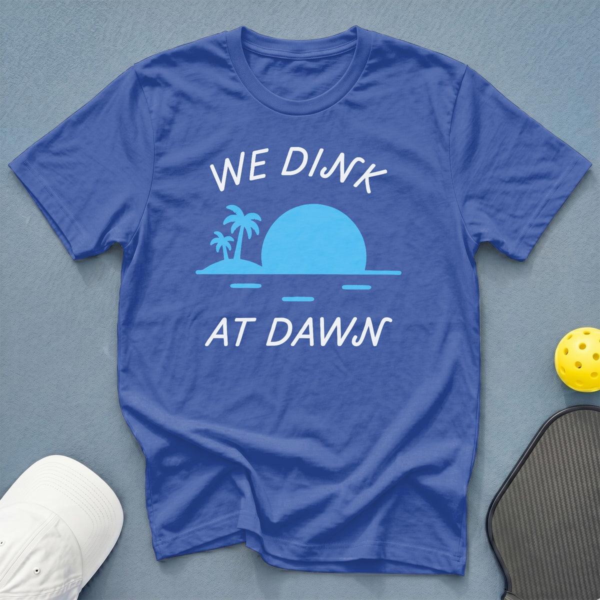 Blue t-shirt with white text "WE DINK AT DAWN" and tropical sunset graphic.