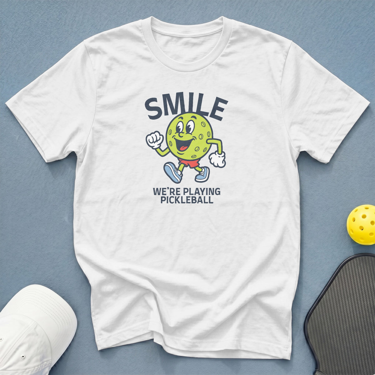 White t-shirt with a cartoon pickleball character and "SMILE WE'RE PLAYING PICKLEBALL" text.