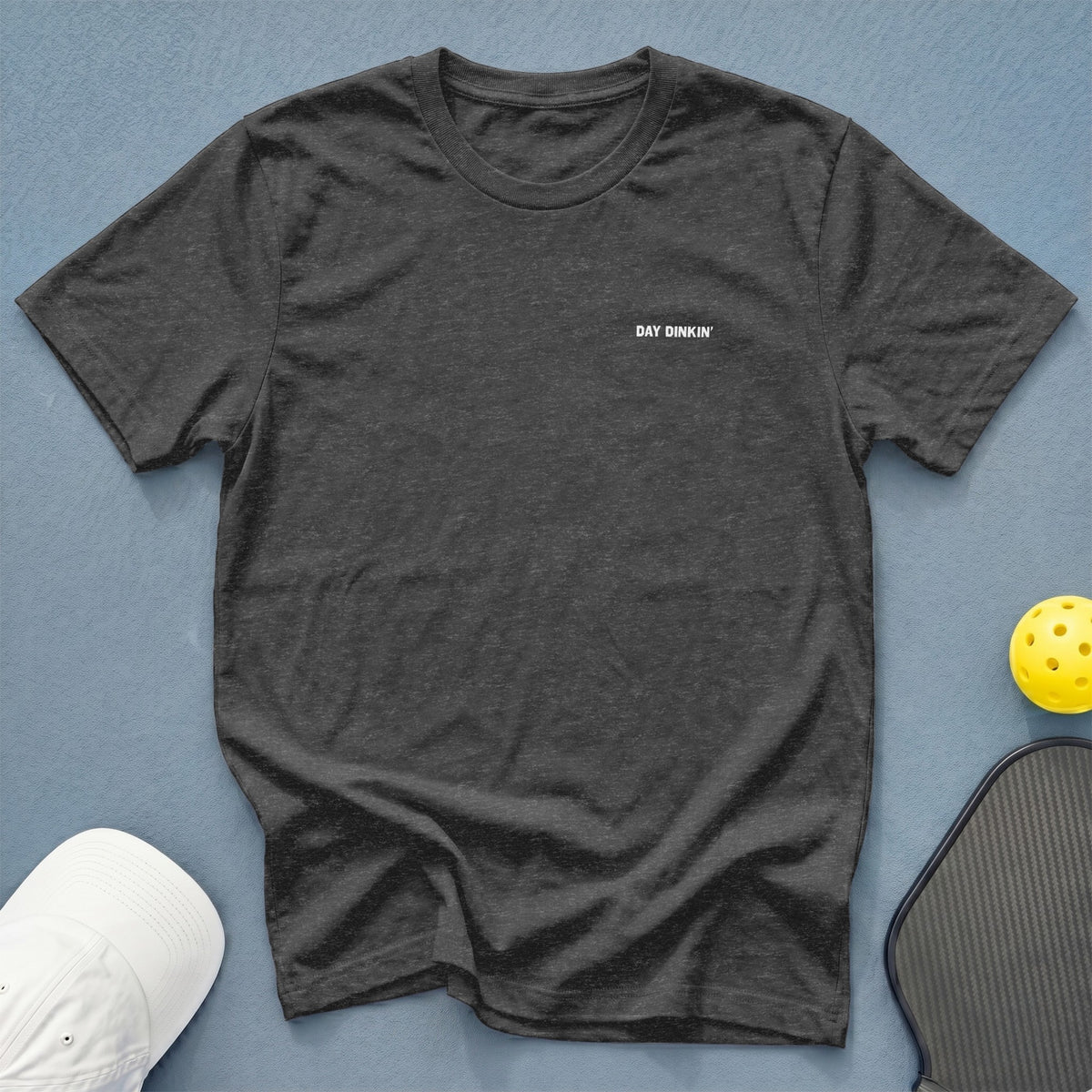 Dark grey t-shirt with white "DAY DINKIN'" text, next to a white cap, pickleball, and paddle.