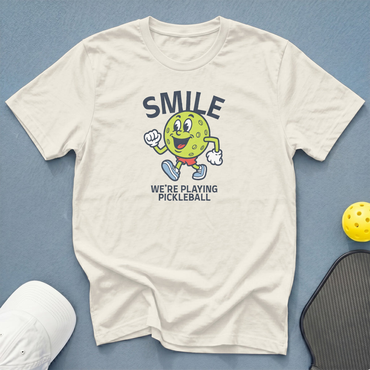 Cream t-shirt with cartoon pickleball, "SMILE" and "WE'RE PLAYING PICKLEBALL" text.