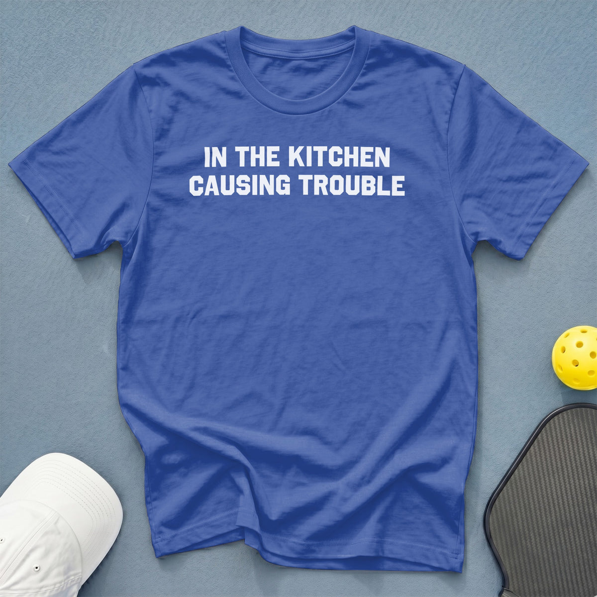Blue t-shirt with white text "IN THE KITCHEN CAUSING TROUBLE".