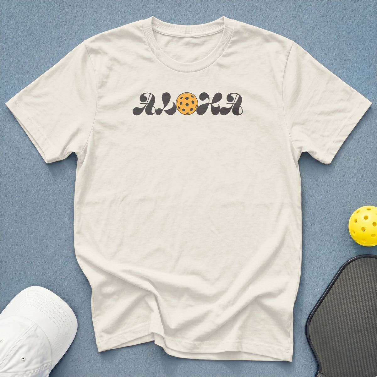 Cream t-shirt with "ALOHA" text and pickleball graphic, cap, and paddle.