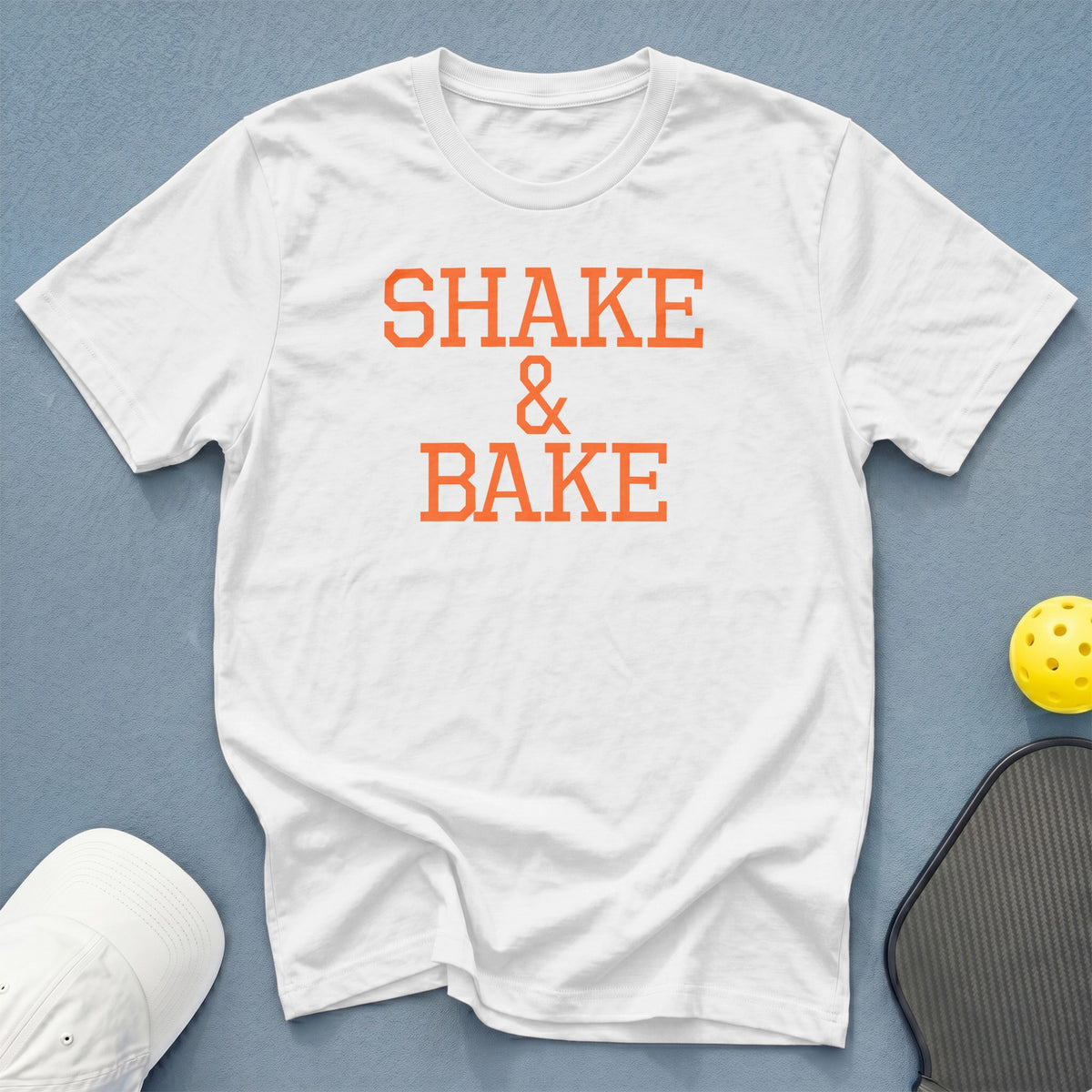 White t-shirt with orange "SHAKE & BAKE" text, pickleball paddle, and ball.