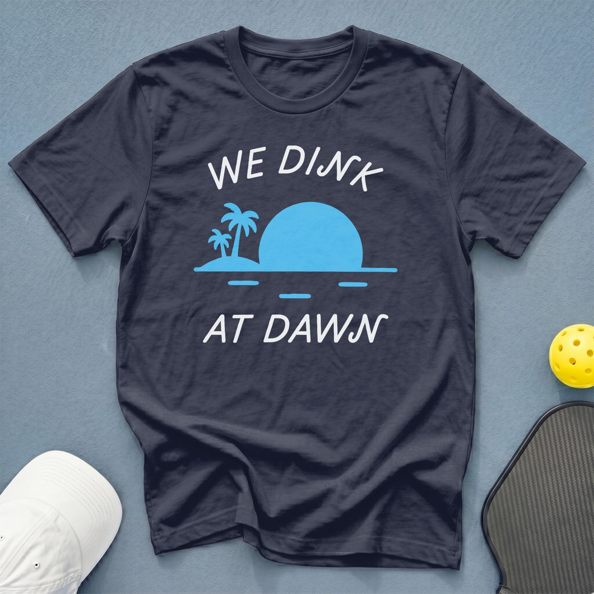 Dark blue t-shirt with "WE DINK AT DAWN" and a sunset beach graphic.