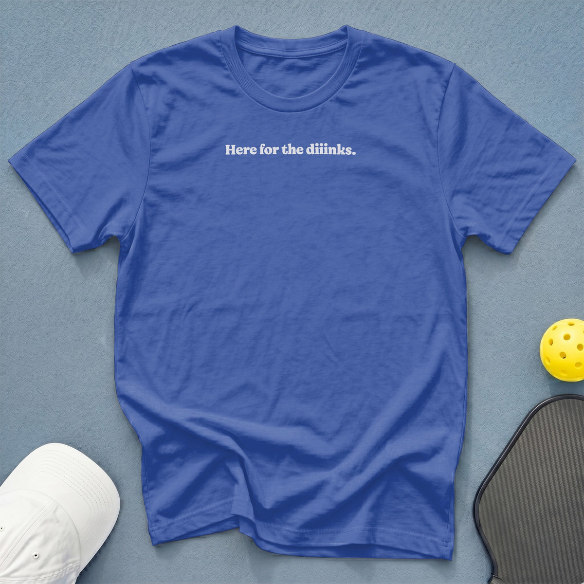 Royal blue t-shirt with white text "Here for the diinks." next to a pickleball paddle and ball.