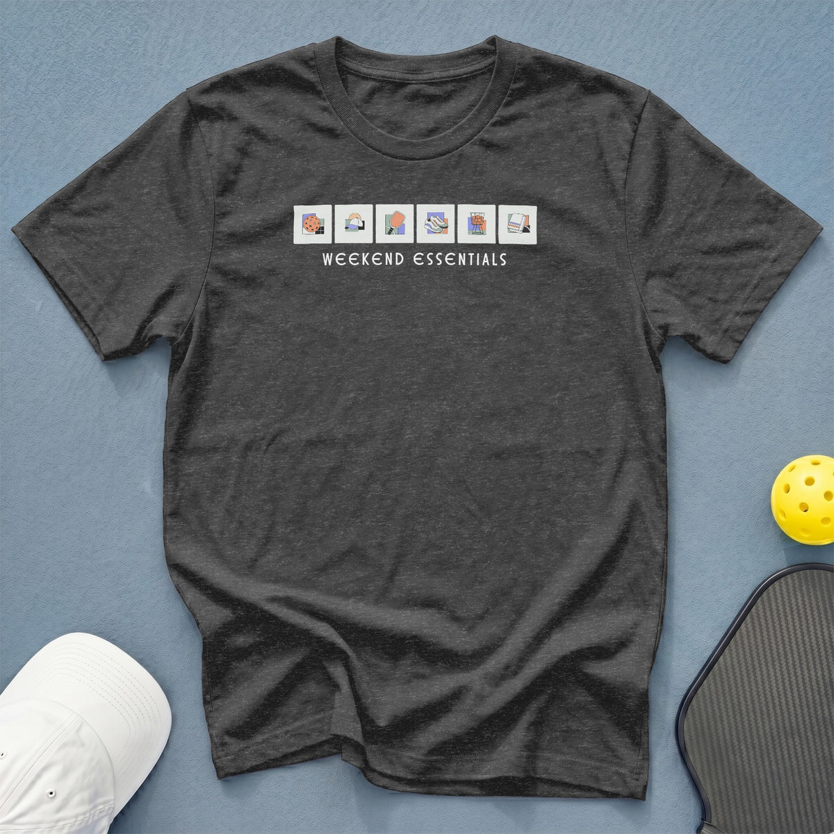 Dark grey t-shirt with "WEEKEND ESSENTIALS" and icons of a cookie, hat, pickleball paddle, shoes, grill, and towel.