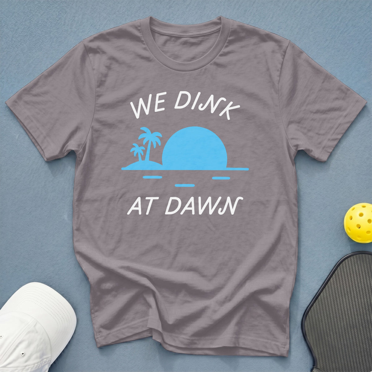Grey t-shirt with "WE DINK AT DAWN" text and palm trees at sunrise graphic.