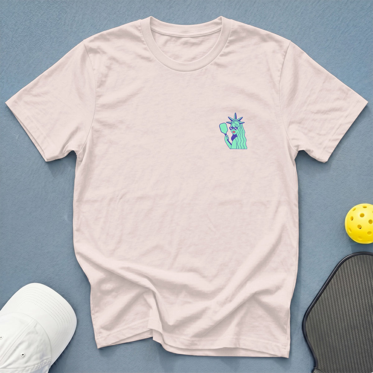 Pale pink t-shirt with a graphic of the Statue of Liberty holding a pickleball paddle.
