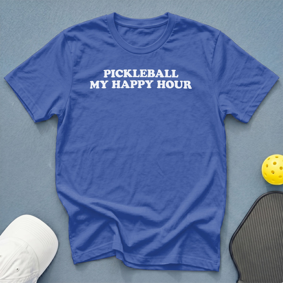 Blue t-shirt with "Pickleball My Happy Hour" text, next to pickleball gear.