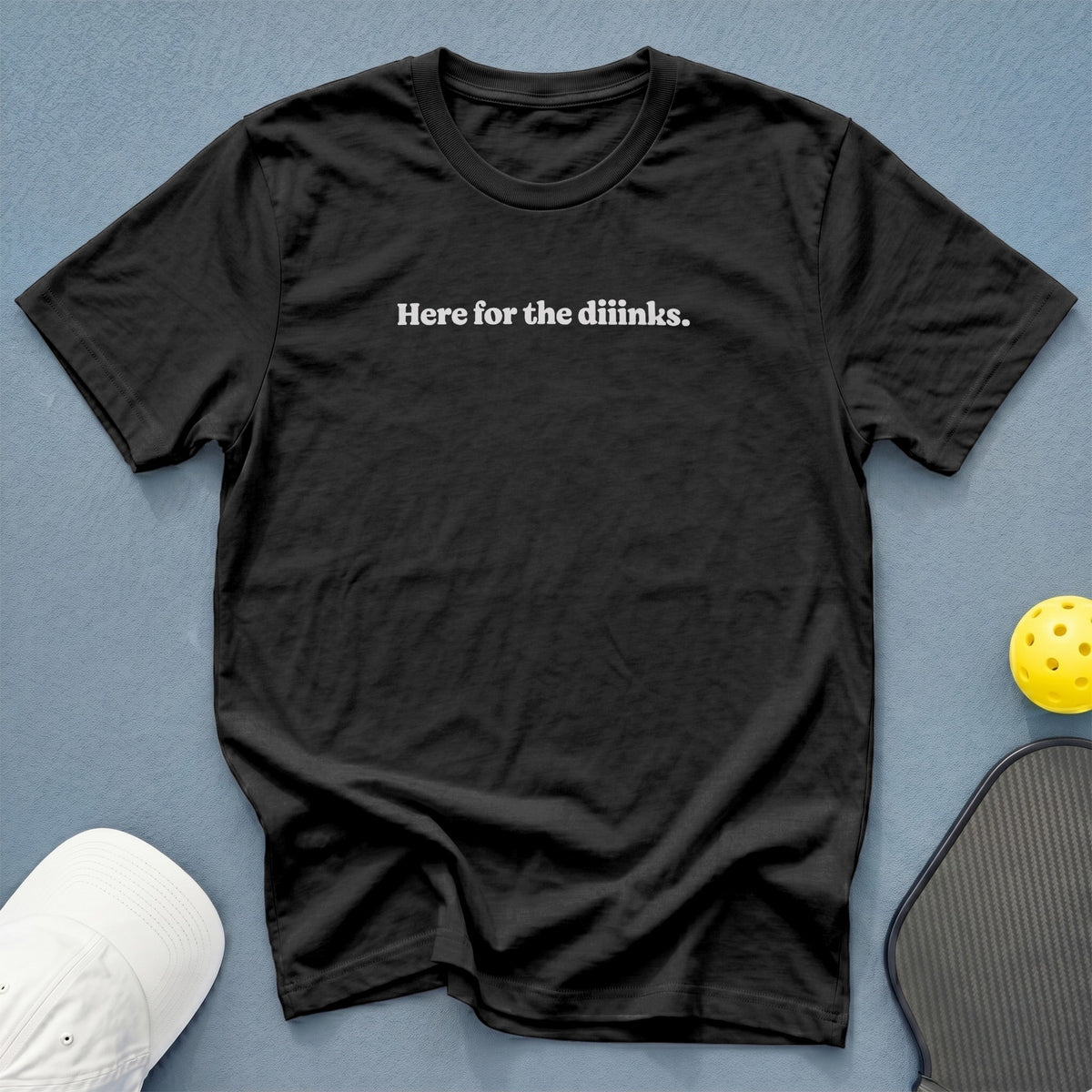 Black t-shirt with white text "Here for the diinks." next to pickleball gear.
