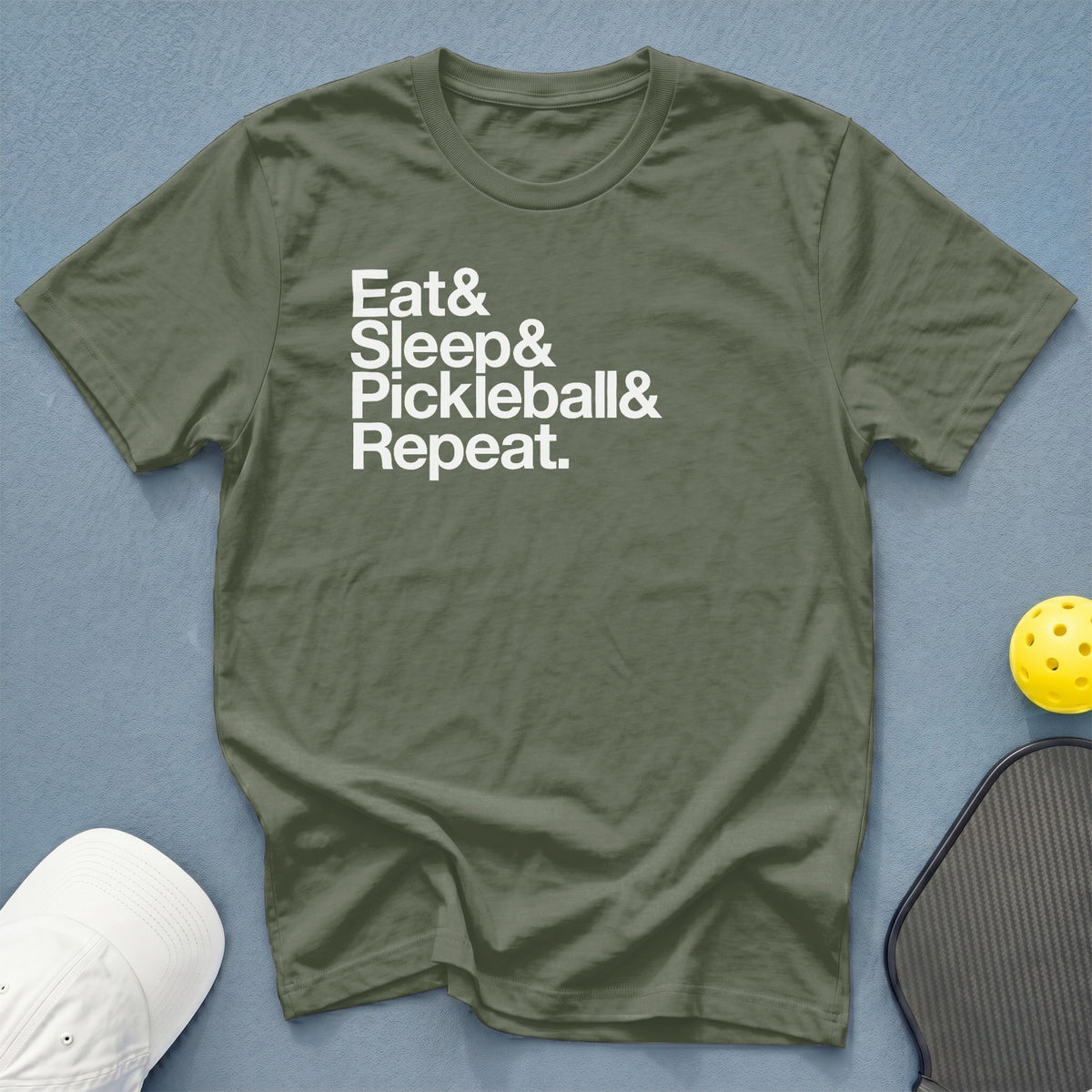 An olive green t-shirt with the white text "Eat& Sleep& Pickleball& Repeat." on a blue textured background, with a white cap, pickleball paddle, and yellow ball nearby.