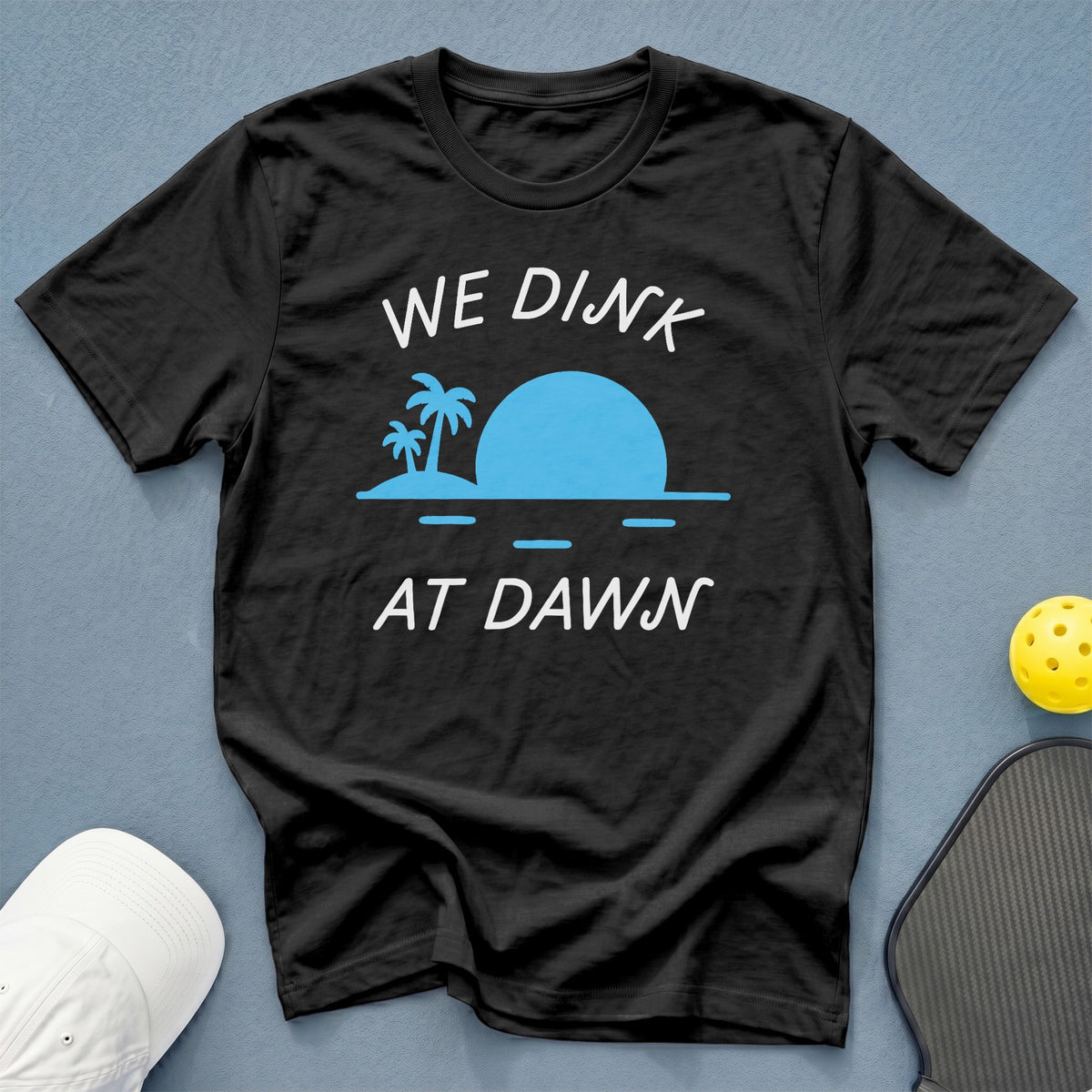Black t-shirt with "WE DINK AT DAWN" and beach sunset graphic.