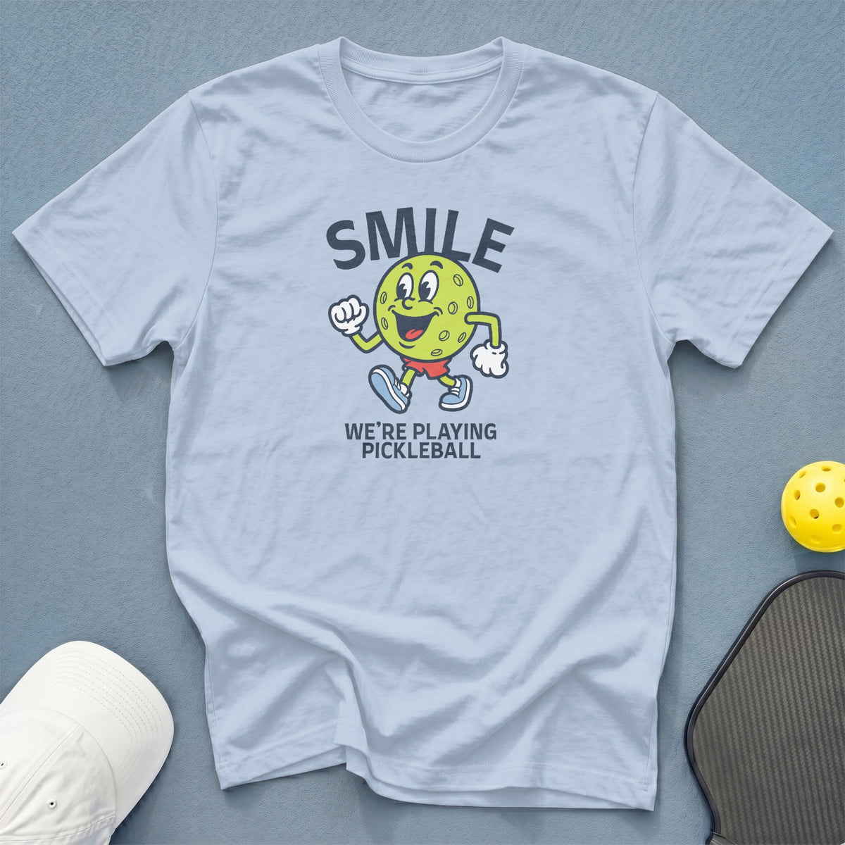 Light blue t-shirt with cartoon pickleball character and text "SMILE WE'RE PLAYING PICKLEBALL".