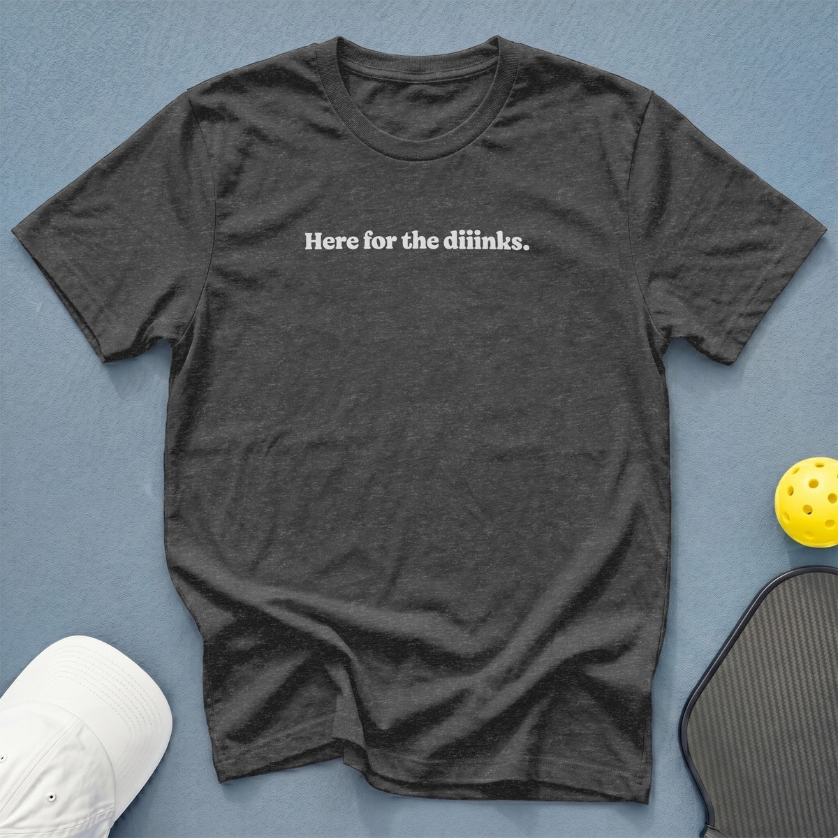 Dark grey t-shirt with "Here for the diiinks." text, white cap, pickleball paddle and ball.
