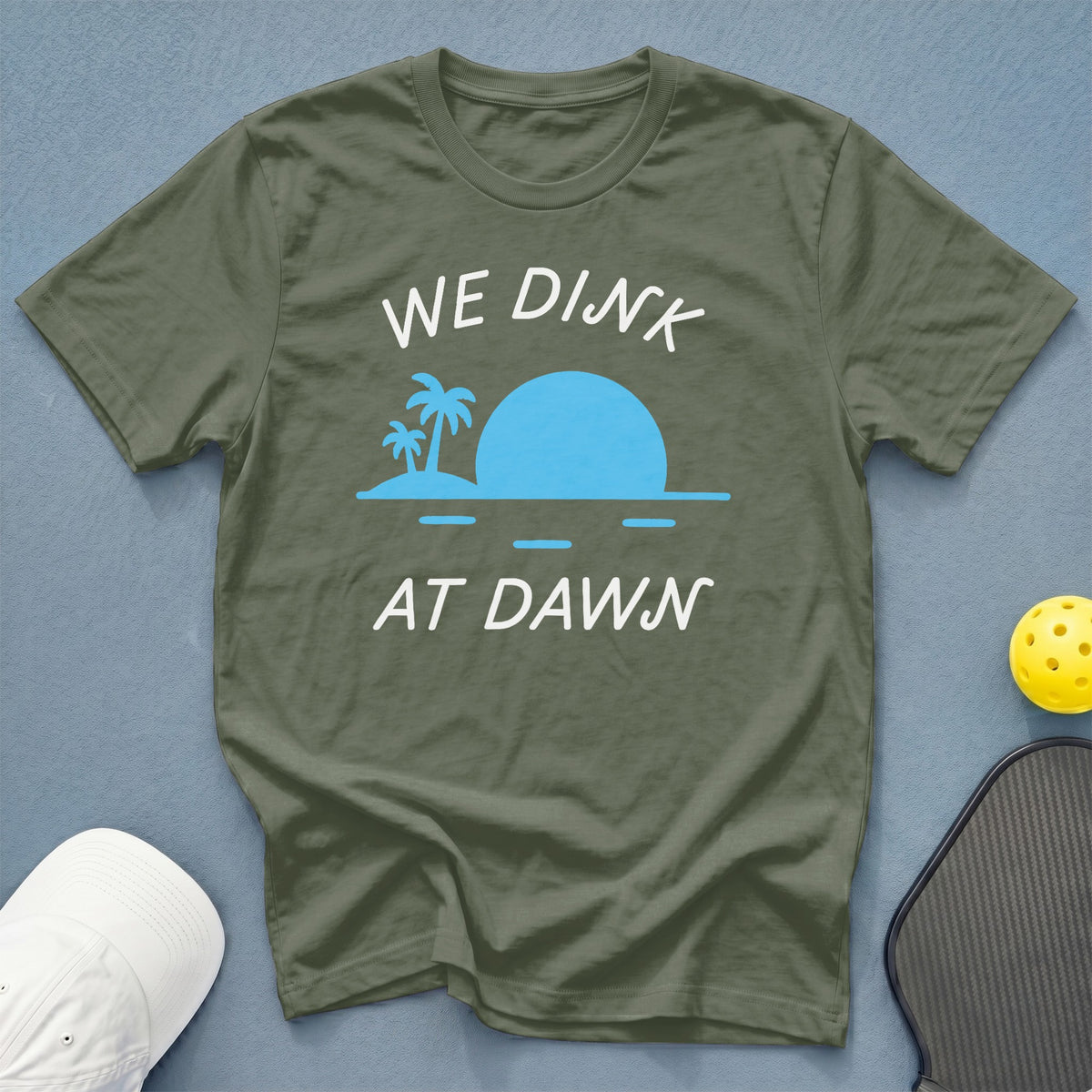 Olive green t-shirt with "WE DINK AT DAWN" text and beach graphic.