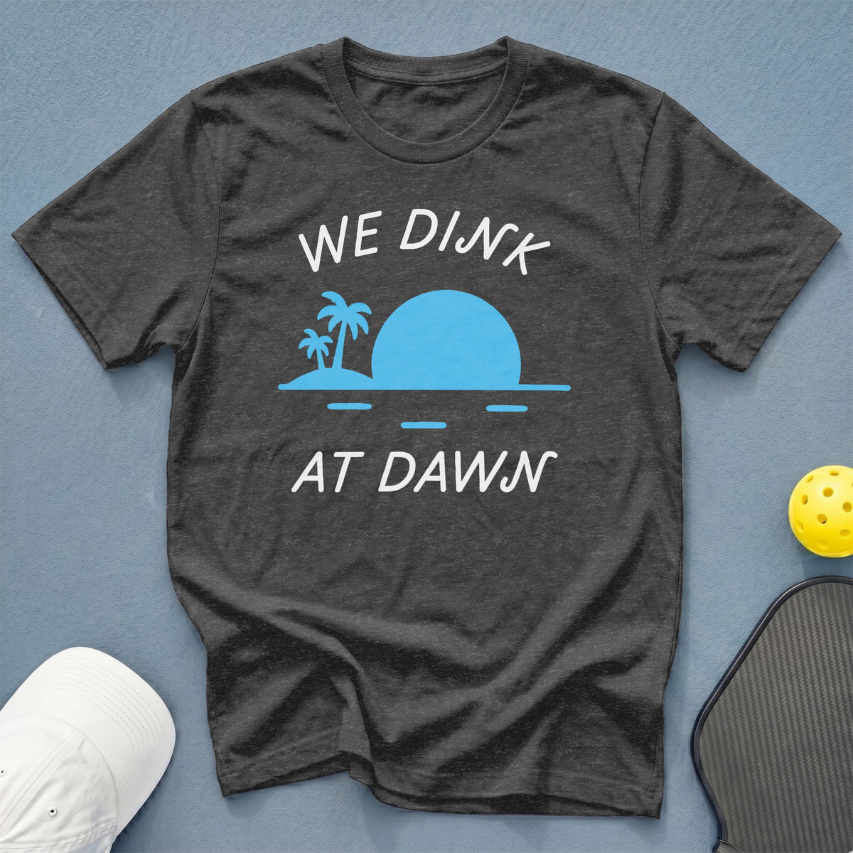 Dark grey t-shirt with "WE DINK AT DAWN" text and tropical sunset graphic.