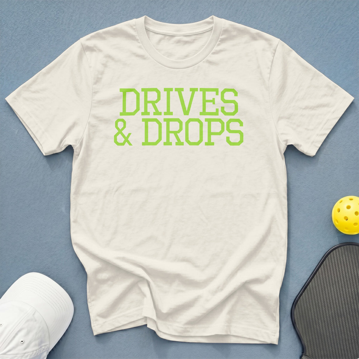 Off-white t-shirt with green "DRIVES & DROPS" text, pickleball paddle, and ball.