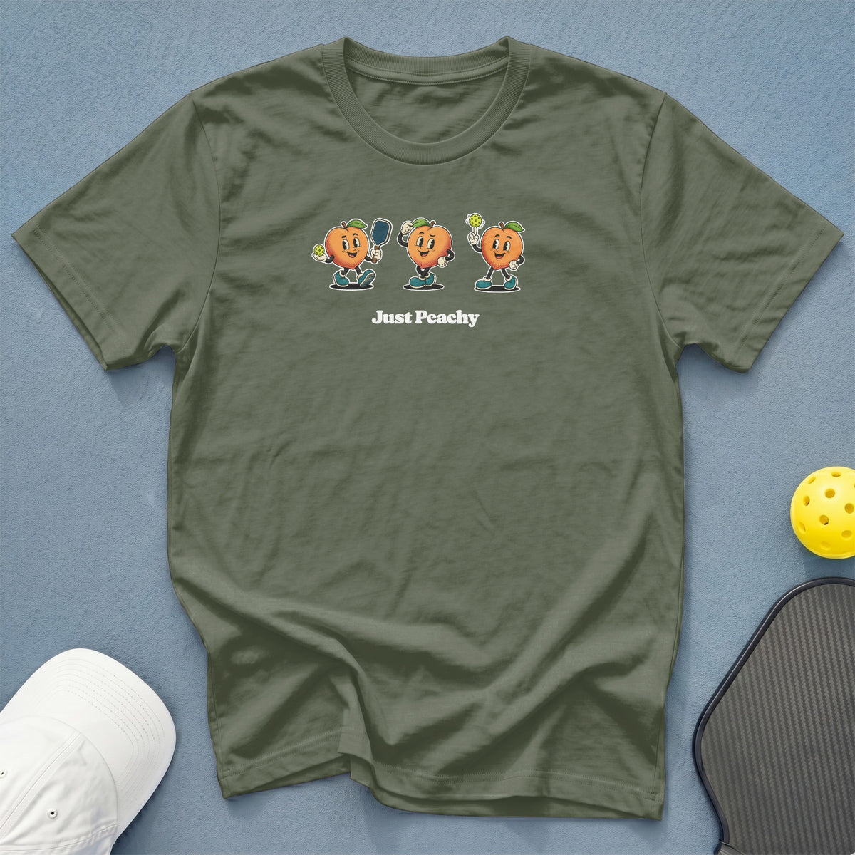 Olive green t-shirt with three cartoon peaches playing pickleball and the text "Just Peachy".