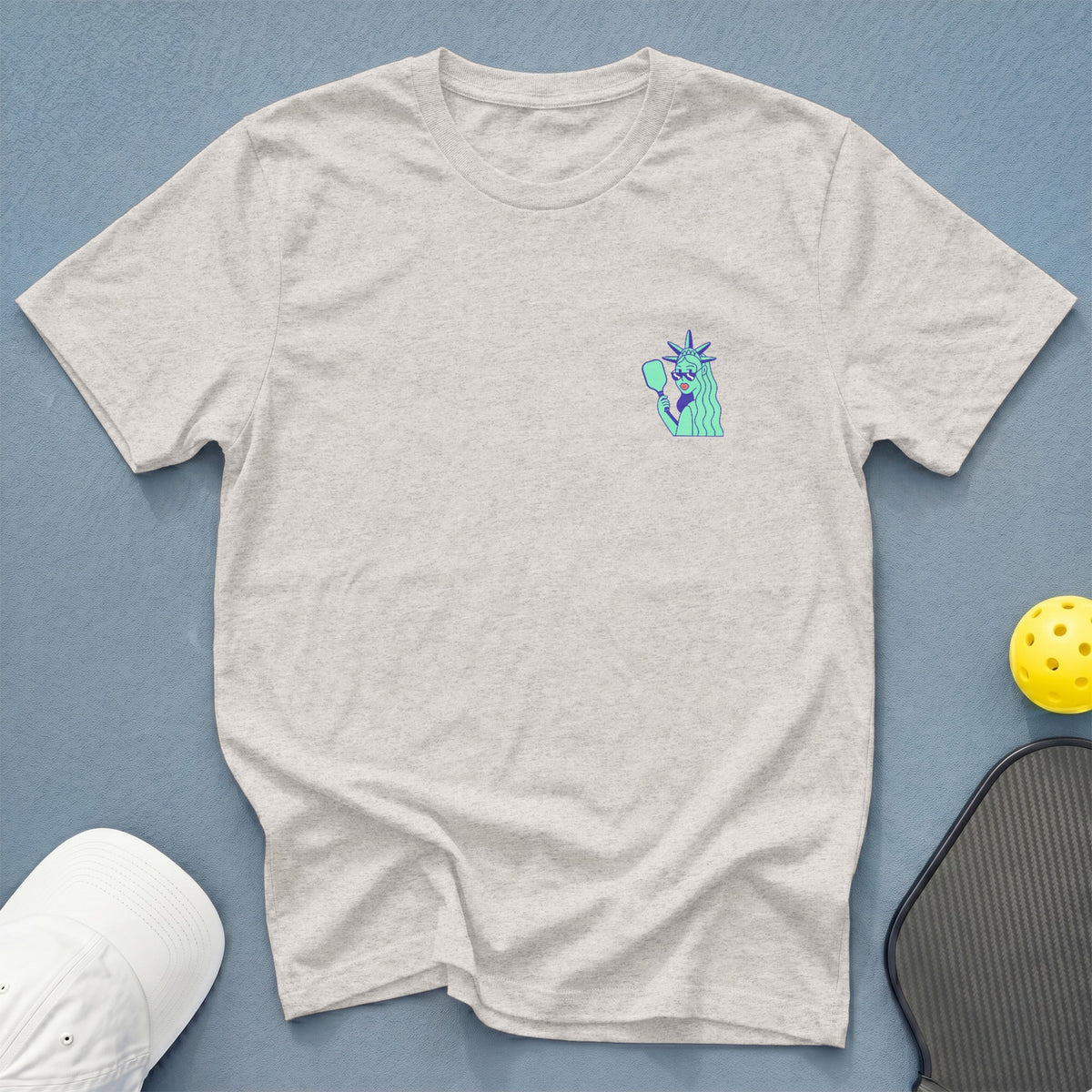 Light gray t-shirt with a blue and green illustration of the Statue of Liberty holding a pickleball paddle.