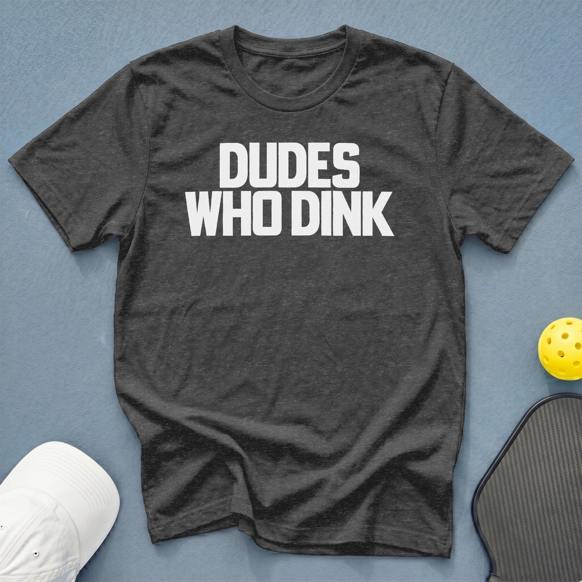 Dark grey t-shirt with white text "DUDES WHO DINK" next to pickleball gear.