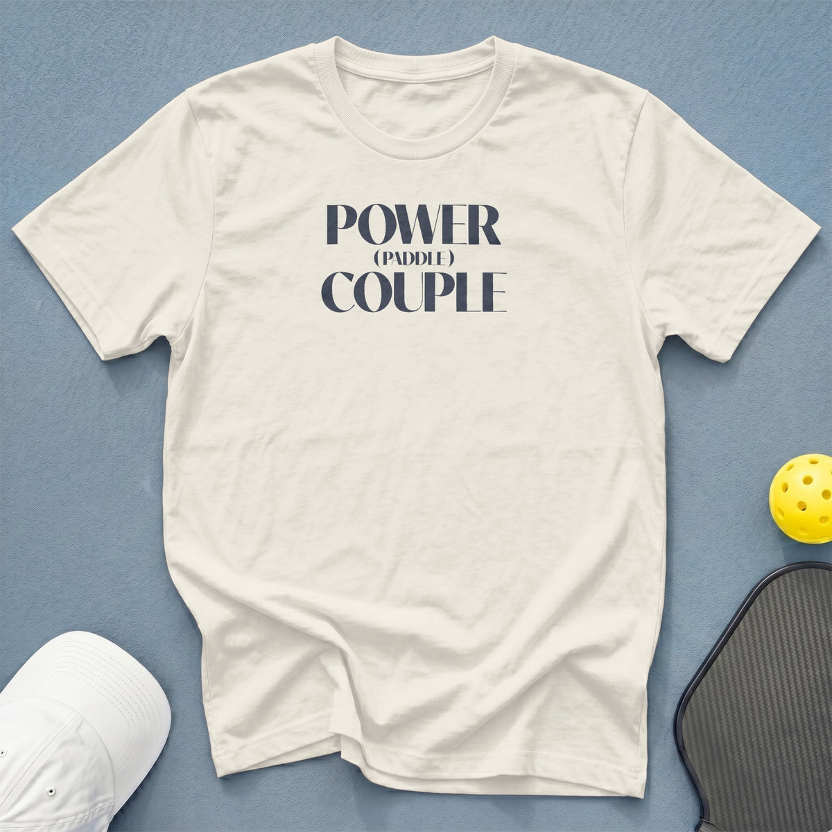 Cream t-shirt with "POWER (PADDLE) COUPLE" text, white hat, pickleball paddle, and ball.