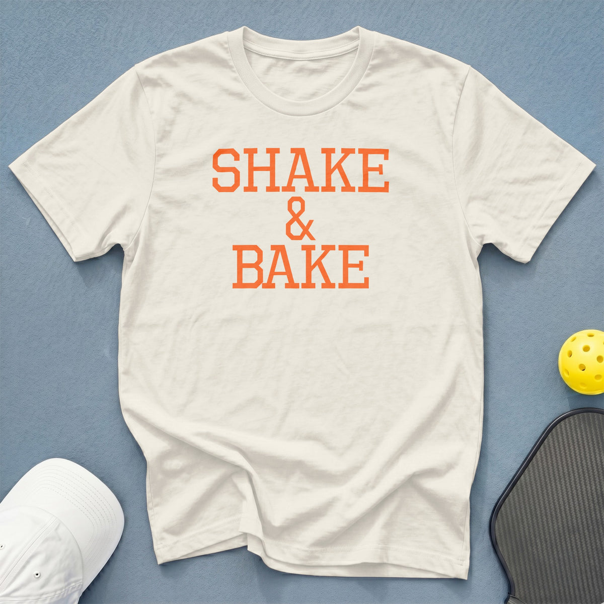 Cream t-shirt with orange text "SHAKE & BAKE", pickleball paddle, and ball.