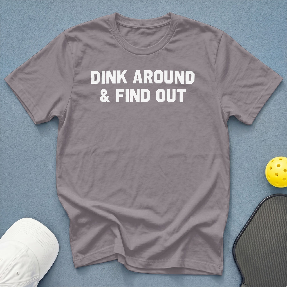 Grey t-shirt with white text "DINK AROUND & FIND OUT", pickleball, and hat.