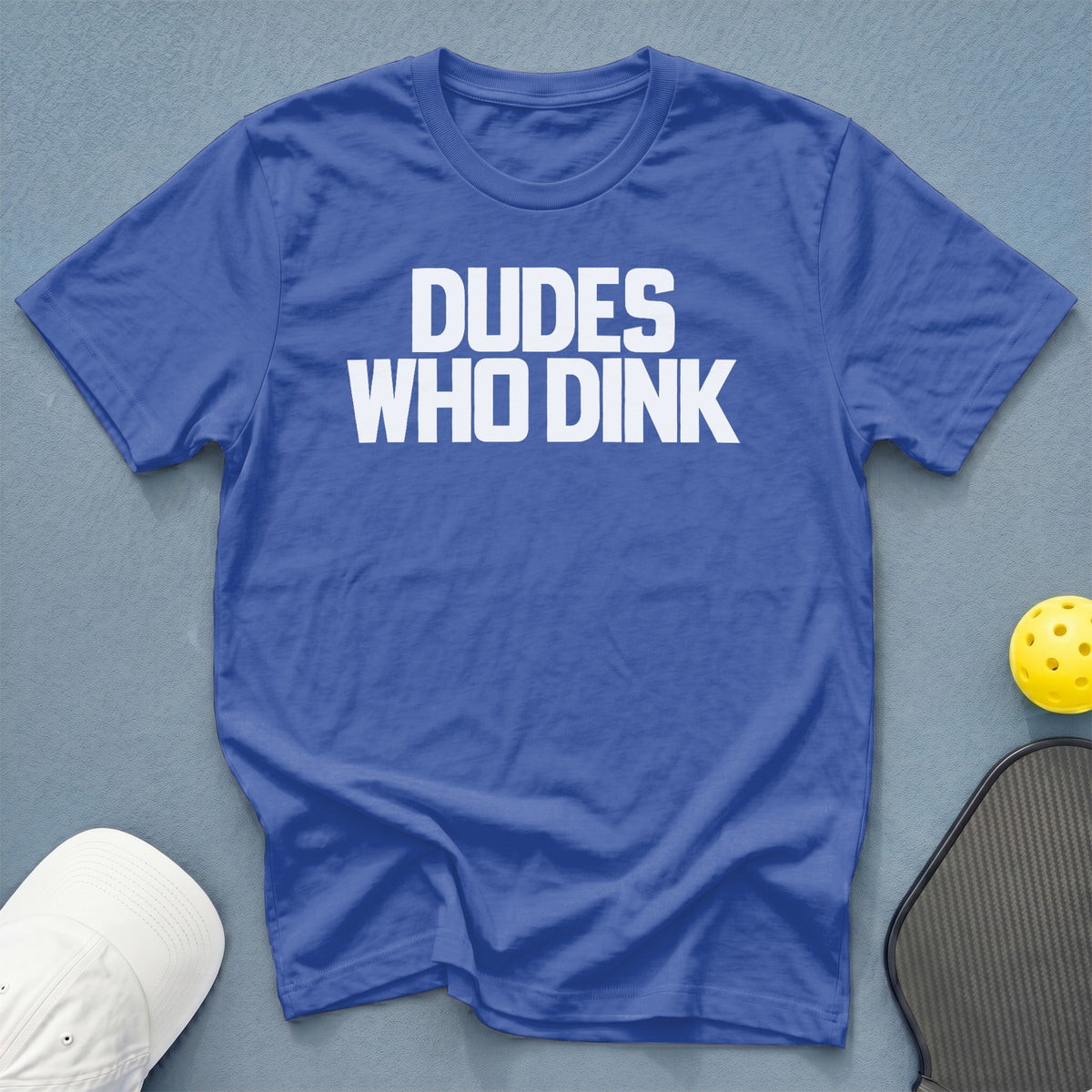 Blue t-shirt with white text reading "Dudes Who Dink", pickleball paddle and ball, white cap.