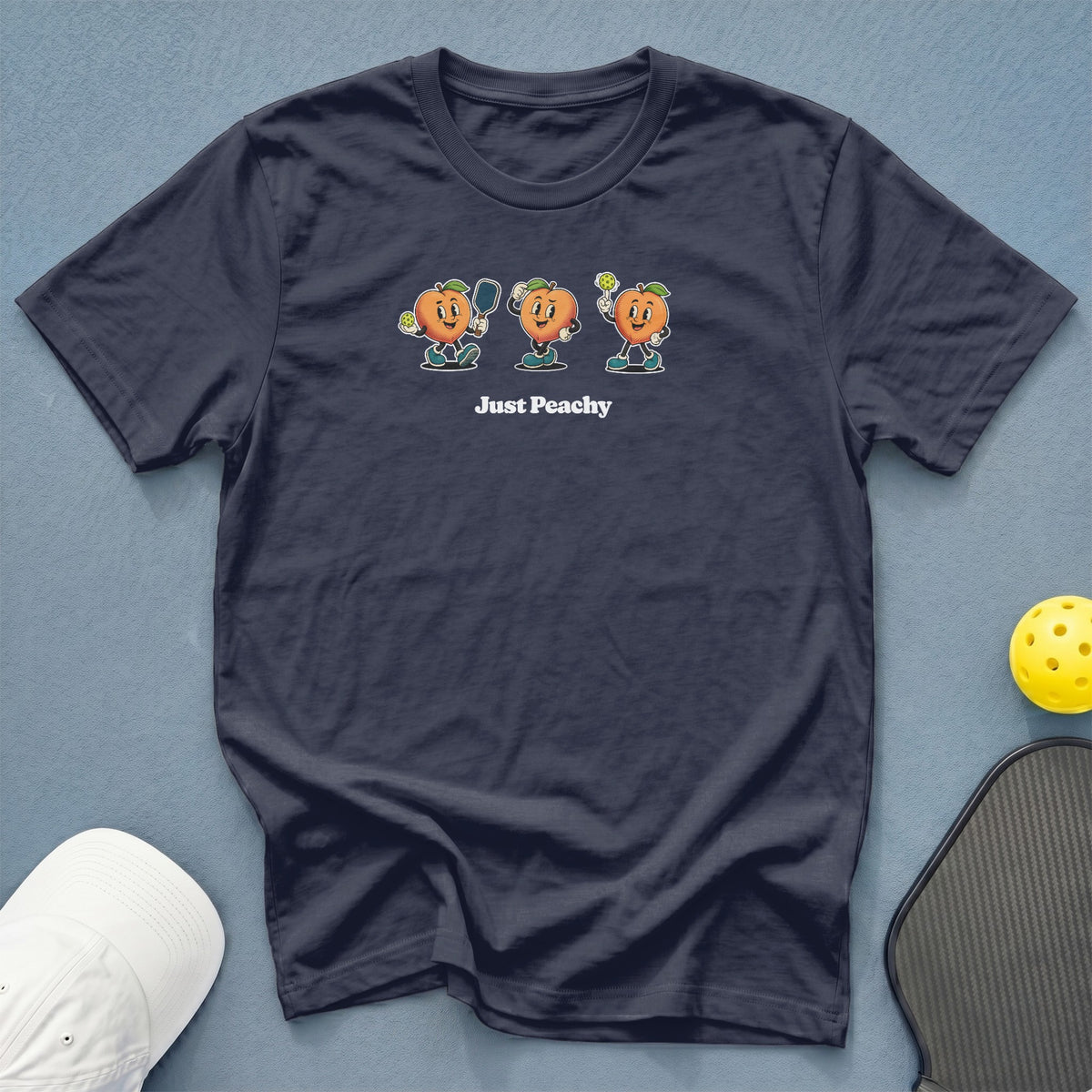 Dark blue t-shirt with cartoon peaches playing pickleball, text "Just Peachy".